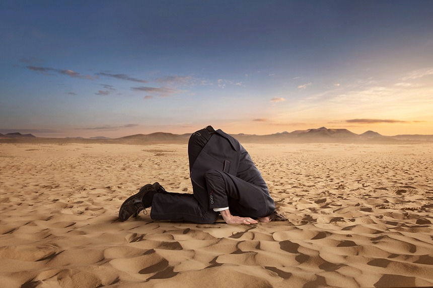 Desperate businessman hiding head in the sand in the desert - We Can No Longer Afford To Ignore Reality! - Miller on the Money