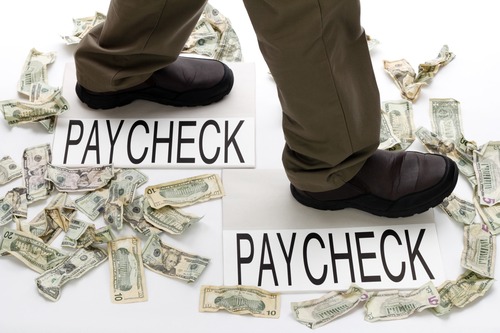 Male legs walking from one paycheck to another with crumpled money scattered about on the floor - You Don't Have To Live From Paycheck To Paycheck! - Miller on the Money